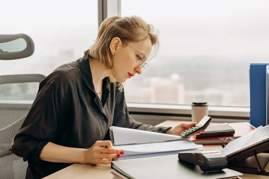 Woman reviewing financial documents and using a calculator at her office desk to check credit score safely.
