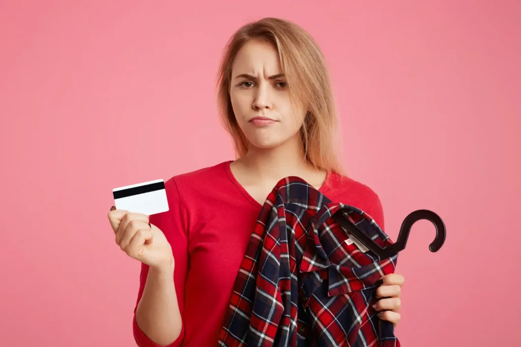 Confused shopper holding store credit card and plaid shirt against pink background, symbolizing store credit card dangers.