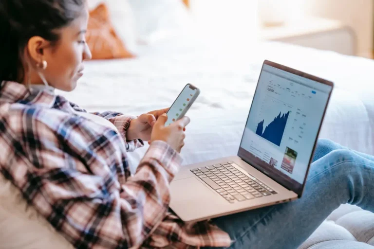 Young woman using smartphone and laptop to track stock market investments online