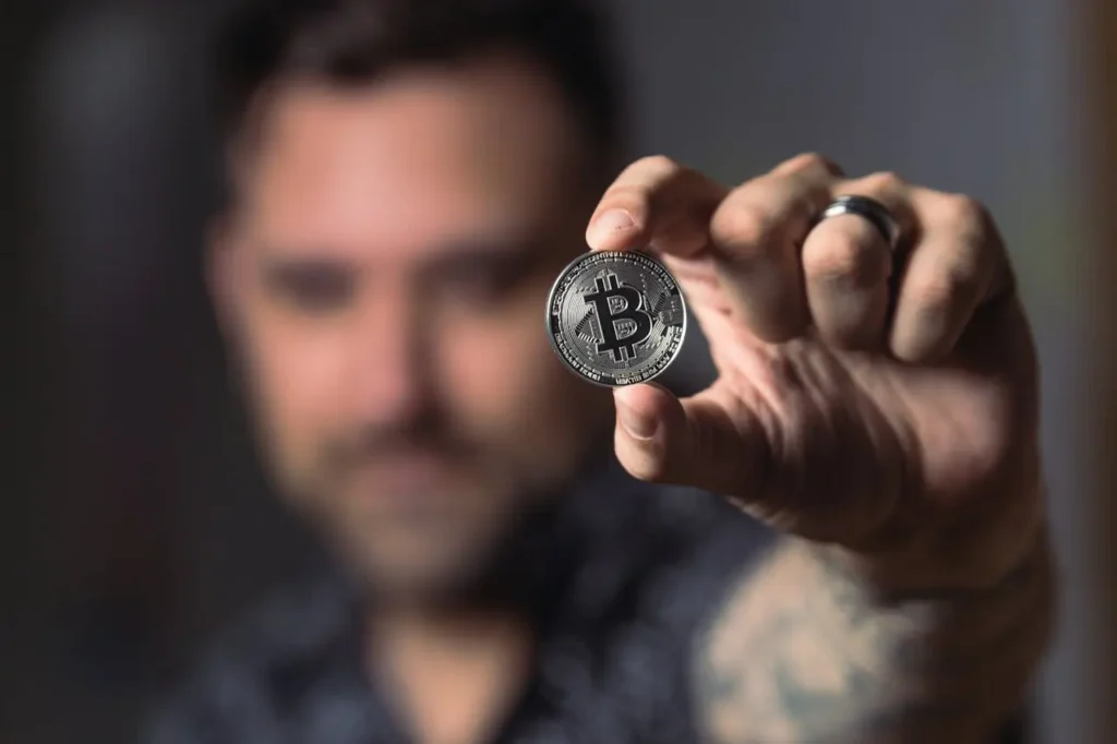 Man holding a silver Bitcoin coin in focus, with blurred background, representing cryptocurrency investing.