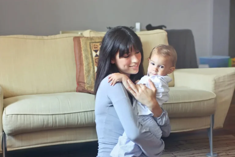 Smiling young woman holding a baby in a cozy living room, representing babysitting services.