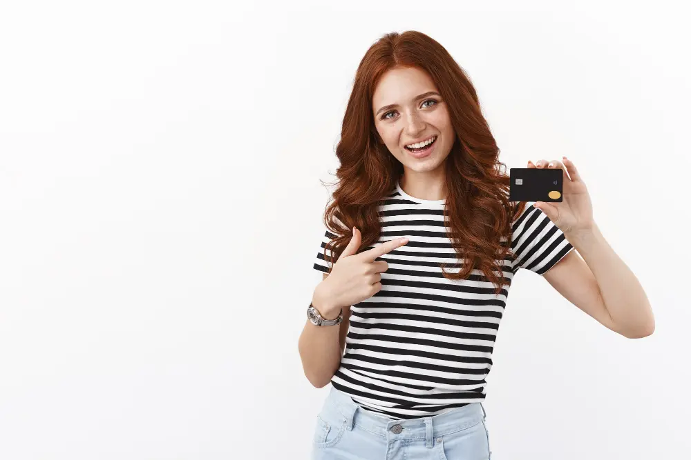 Smiling young woman pointing to her first credit card while standing against a white background