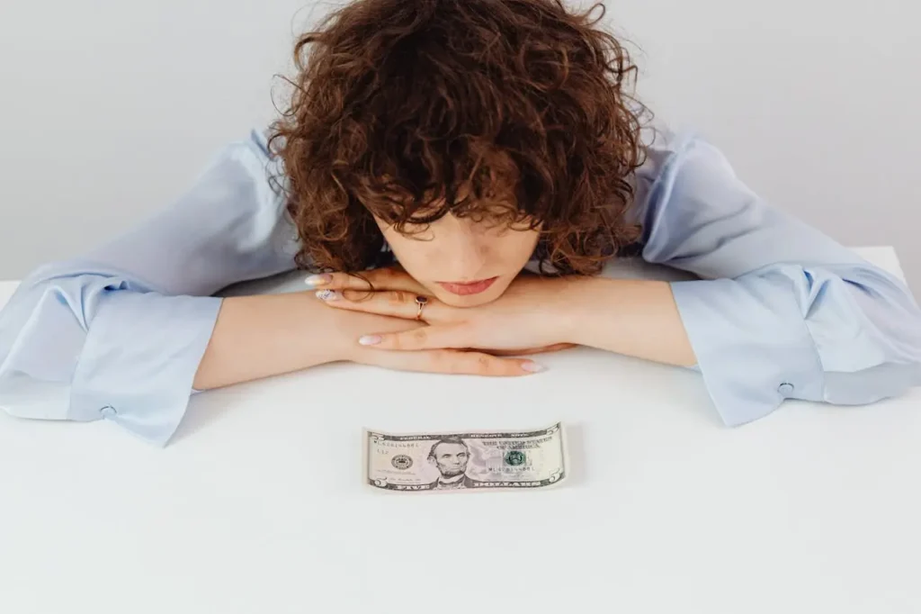 Worried woman looking at a five-dollar bill on a white table, symbolizing financial stress and the dilemma of whether to save or pay off debt first.