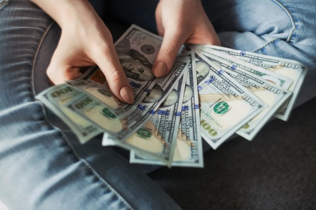 Person holding multiple $100 bills in their hands while sitting cross-legged, symbolizing passive income ideas and financial freedom.