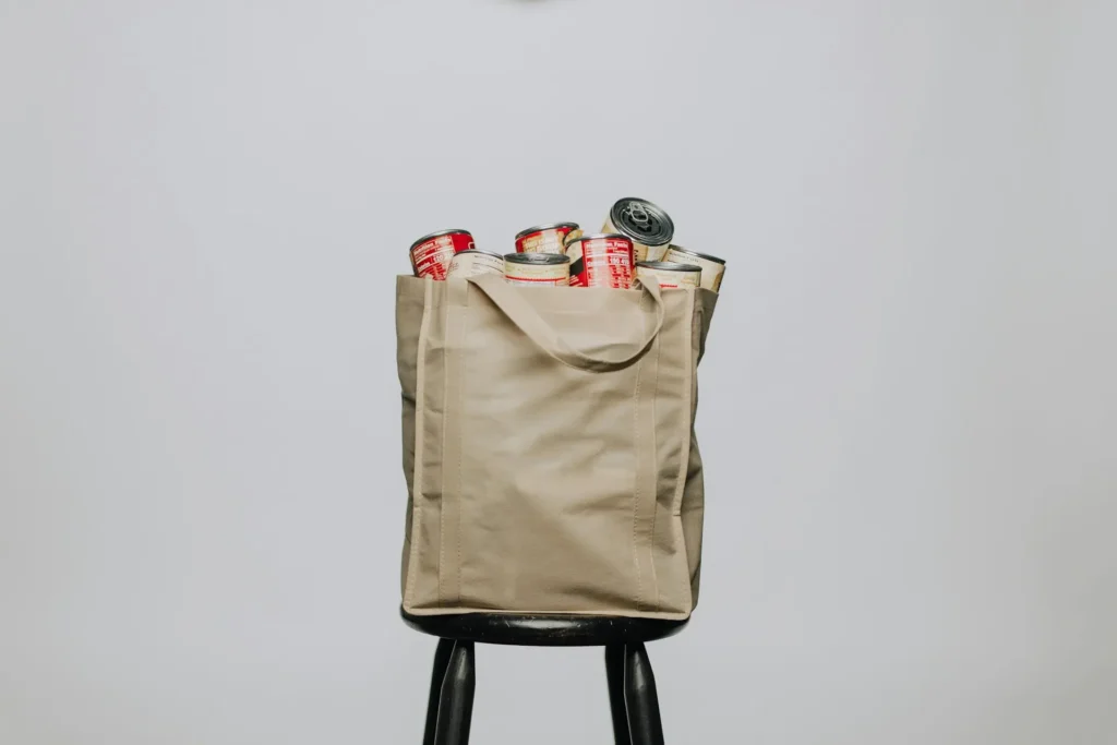 Reusable grocery bag filled with canned goods, representing smart bulk buying strategies, placed on a stool against a neutral background.