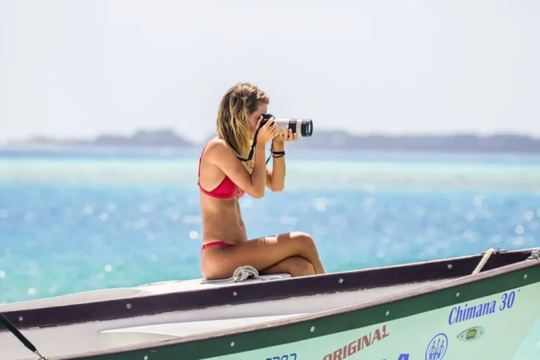 Woman in a red bikini taking photos with a DSLR camera while sitting on a boat in tropical blue waters, representing passive income ideas through photography as a revenue stream.