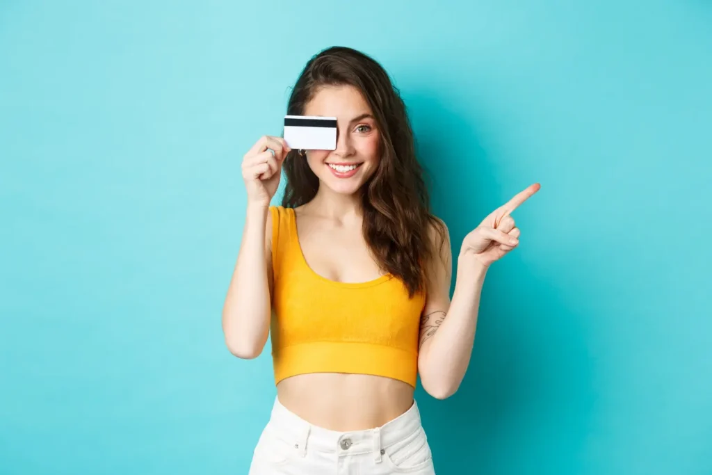 Smiling young woman holding a credit card over one eye and pointing upward against a blue background.