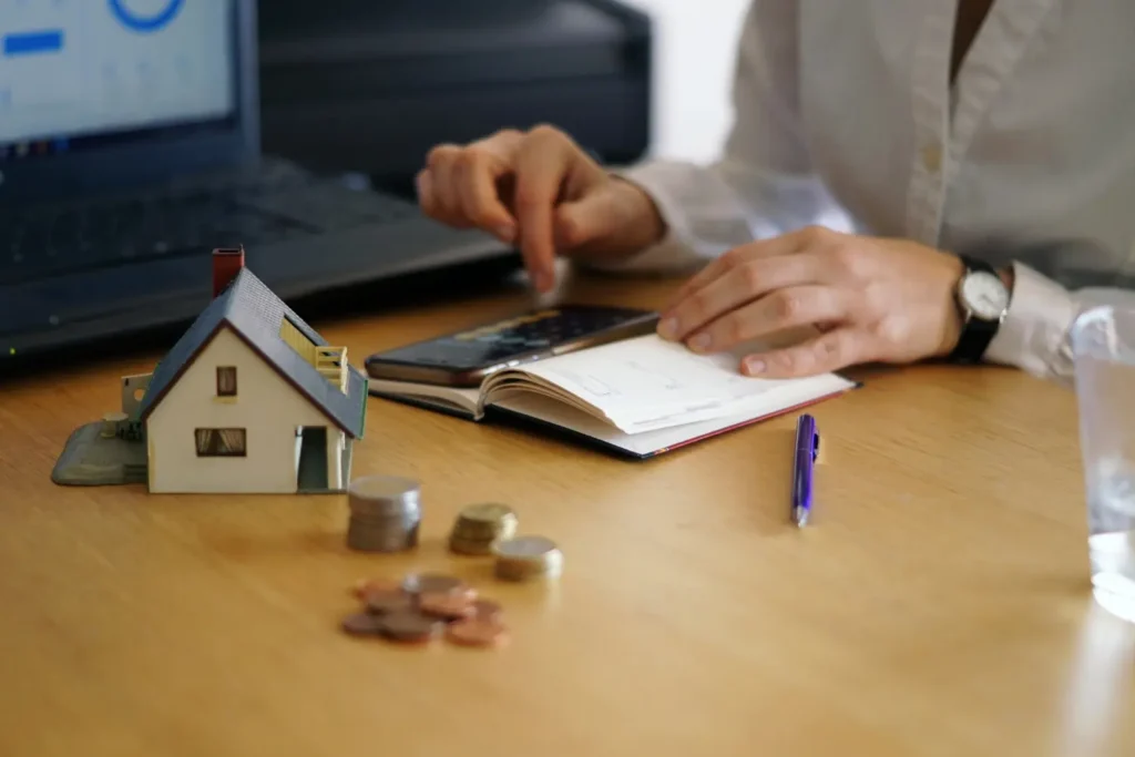 Person calculating 2026 mortgage rates with coins, miniature house model, and financial planner on desk.