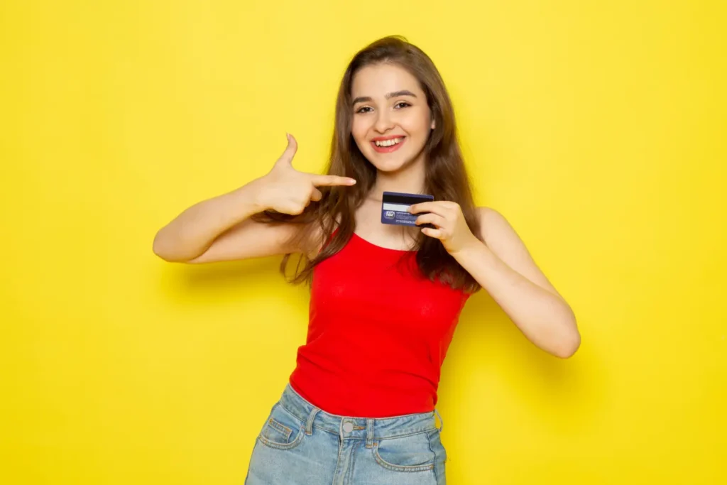 Young woman smiling and holding a credit card against a bright yellow background, symbolizing how to build credit with a credit card responsibly.